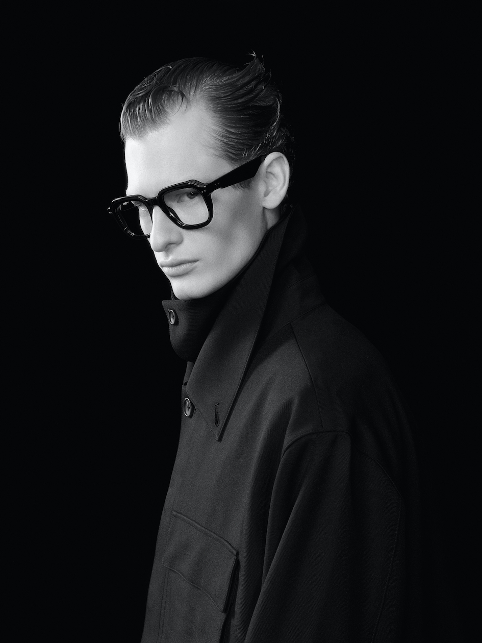 Side view of a model in a dark, minimalist coat wearing the Takumi frames, showcasing the bold silhouette and the contrast between the acetate and industrial metal elements.