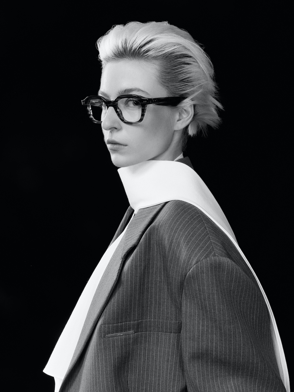Profil en noir et blanc d'un mannequin portant les lunettes Fumaki en acétate travaillé, accompagnées d'un manteau architectural à col haut.