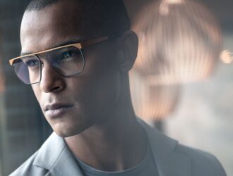 Close-up of a man wearing the Sandpoint frames, featuring a squared design with a prominent gold double bridge and blue-rimmed lenses.