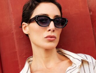 Femme portant des lunettes de soleil œil-de-chat noires avec détails en strass sur fond de porte rouge