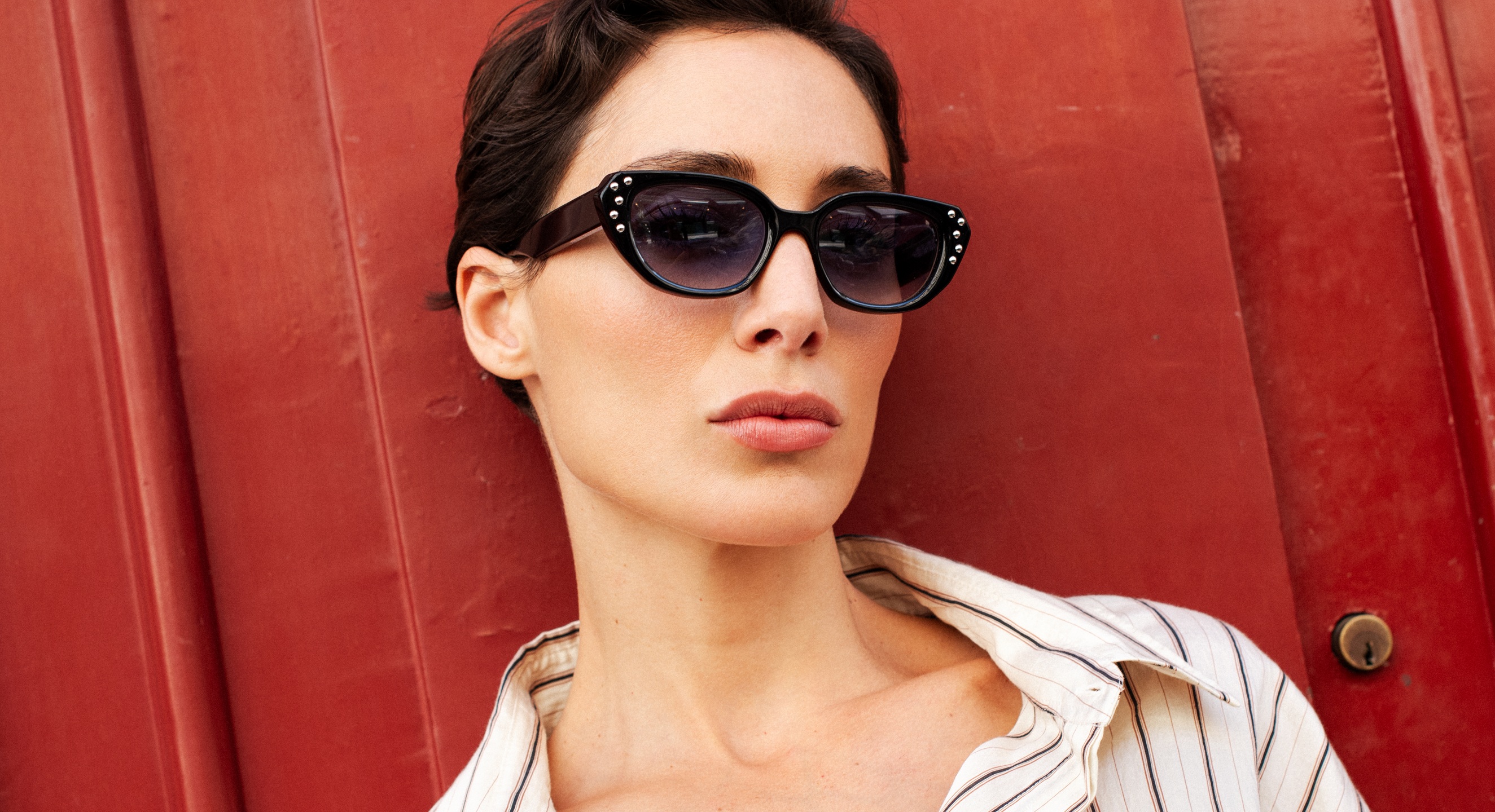 Femme portant des lunettes de soleil œil-de-chat noires avec détails en strass sur fond de porte rouge