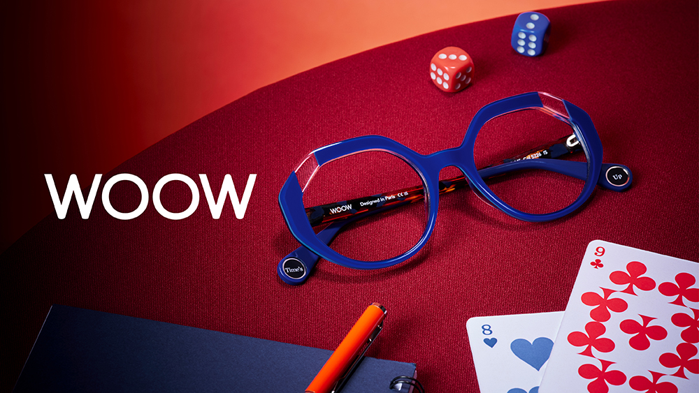 Visuel de campagne WOOW présentant les lunettes optiques Times Up 2 en bleu cobalt sur une surface rouge, entourées de cartes à jouer, de dés et d'un stylo