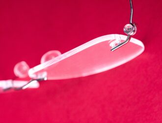 Close-up detail of a rimless eyeglass lens with pressure-fit mounting system against a red background.