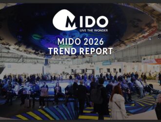 Large exhibition hall at MIDO 2026 eyewear trade show with attendees gathered around a circular blue-lit stage area. The MIDO logo with tagline 'Live the Wonder' and text 'MIDO 2026 TREND REPORT' overlays the scene. Exhibition booths from brands like Optovisol, Desio, and Visionix visible in the background.