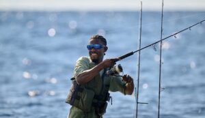 Angler Emmanuel Williams smiles while holding a fishing rod on the open water, wearing the blue-lensed Pilothouse PRO shield sunglasses.