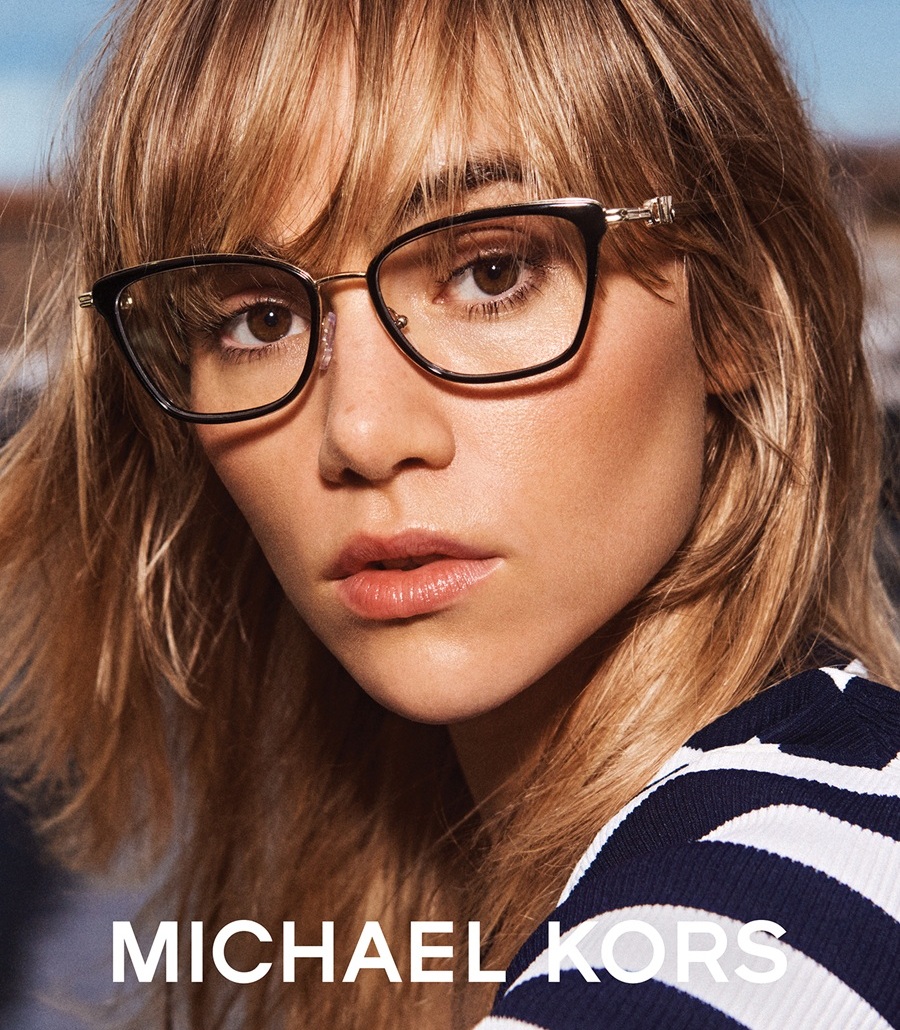 Un gros plan de Suki Waterhouse portant des lunettes de vue papillon noires et un haut à rayures marines et blanches, avec le logo Michael Kors en bas.