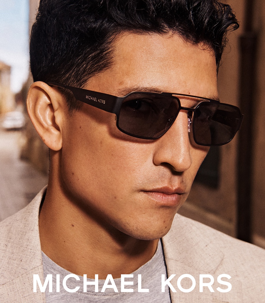 Un gros plan de Danny Ramirez de profil, arborant des lunettes de soleil Michael Kors foncées et rectangulaires avec un blazer en lin beige.