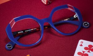 Close-up of WOOW Times Up 2 optical glasses in deep blue with hexagonal frames and tortoiseshell temples, resting on a red surface beside a playing card
