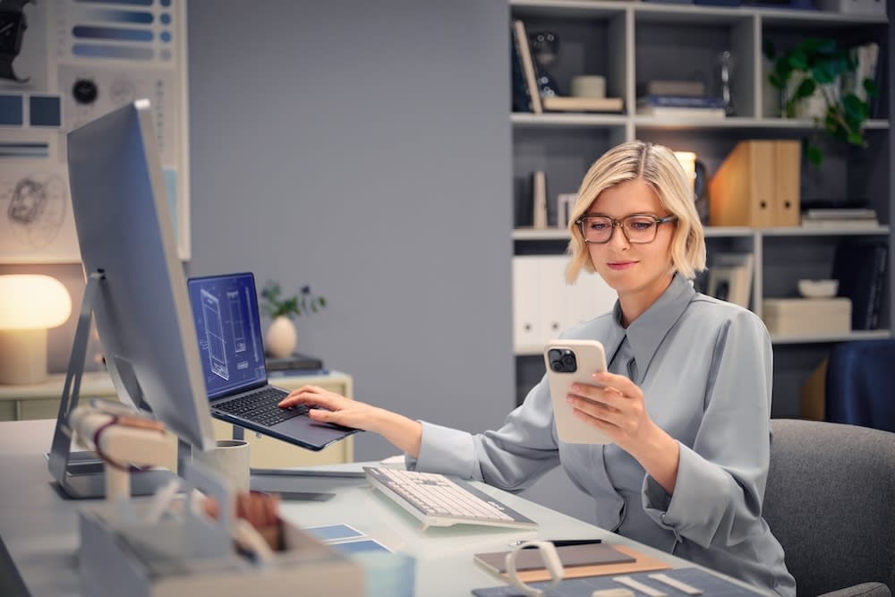 Une femme professionnelle portant des lunettes à son bureau, alternant entre un grand écran, un ordinateur portable et un smartphone pour illustrer le champ de vision de proximité des verres Varilux Immersia.
