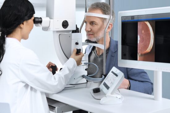 A clinician using the ZEISS VISULAS yag therapeutic laser to treat a male patient, with a monitor beside them displaying a close-up retinal image captured during the procedure.