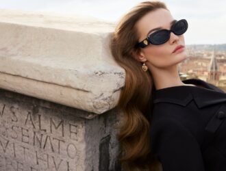 A model reclines against an ancient Roman stone monument inscribed with Latin text, wearing Dolce&Gabbana DG4537 black oval sunglasses with a gold Sacred Heart temple detail, dressed in a structured black coat, with the Rome skyline visible in the background. SS26 Devotion Eyewear campaign.