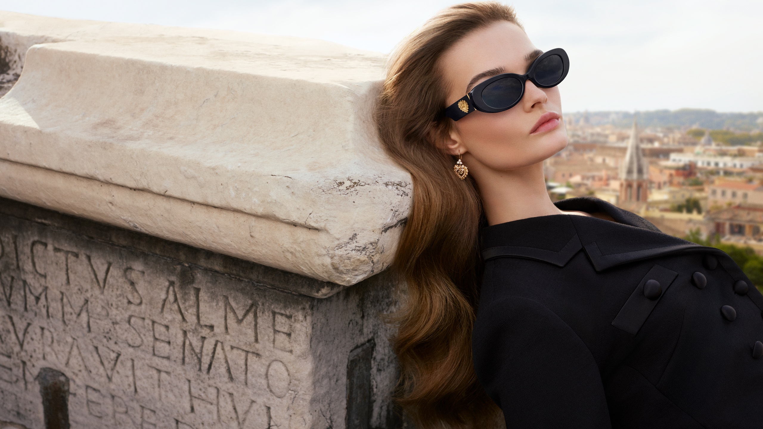 A model reclines against an ancient Roman stone monument inscribed with Latin text, wearing Dolce&Gabbana DG4537 black oval sunglasses with a gold Sacred Heart temple detail, dressed in a structured black coat, with the Rome skyline visible in the background. SS26 Devotion Eyewear campaign.