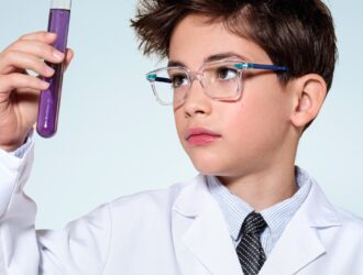A boy in a white lab coat and tie holds up a test tube filled with purple liquid and examines it with focused curiosity, wearing clear-framed glasses with blue arms — portraying the scientist of tomorrow.