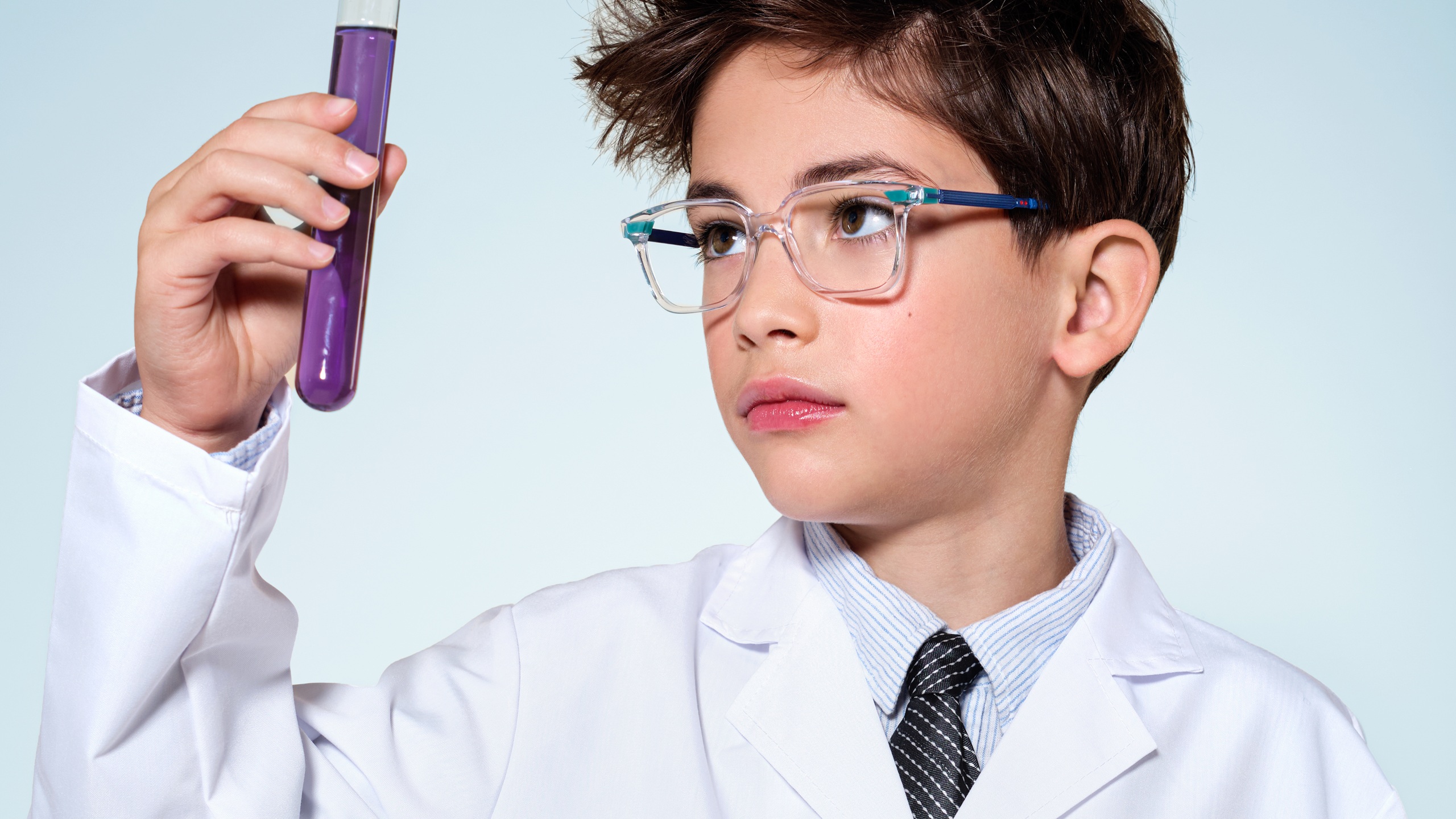 A boy in a white lab coat and tie holds up a test tube filled with purple liquid and examines it with focused curiosity, wearing clear-framed glasses with blue arms — portraying the scientist of tomorrow.