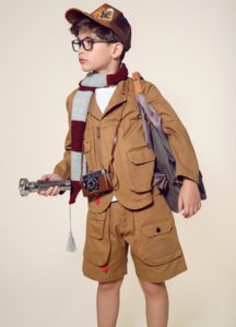 A boy dressed as an adventurer — wearing a khaki jacket and shorts, a patch-covered cap, a striped scarf, and dark-framed glasses — holds a flashlight and carries a backpack and vintage camera, posed as a young explorer ready for discovery.