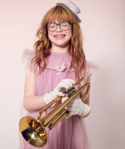 A red-haired girl in a pink tulle dress, lace gloves, a floral hat, and blue glasses beams with joy while holding a brass trumpet, embodying the spirit of a young musician lost in her own world.