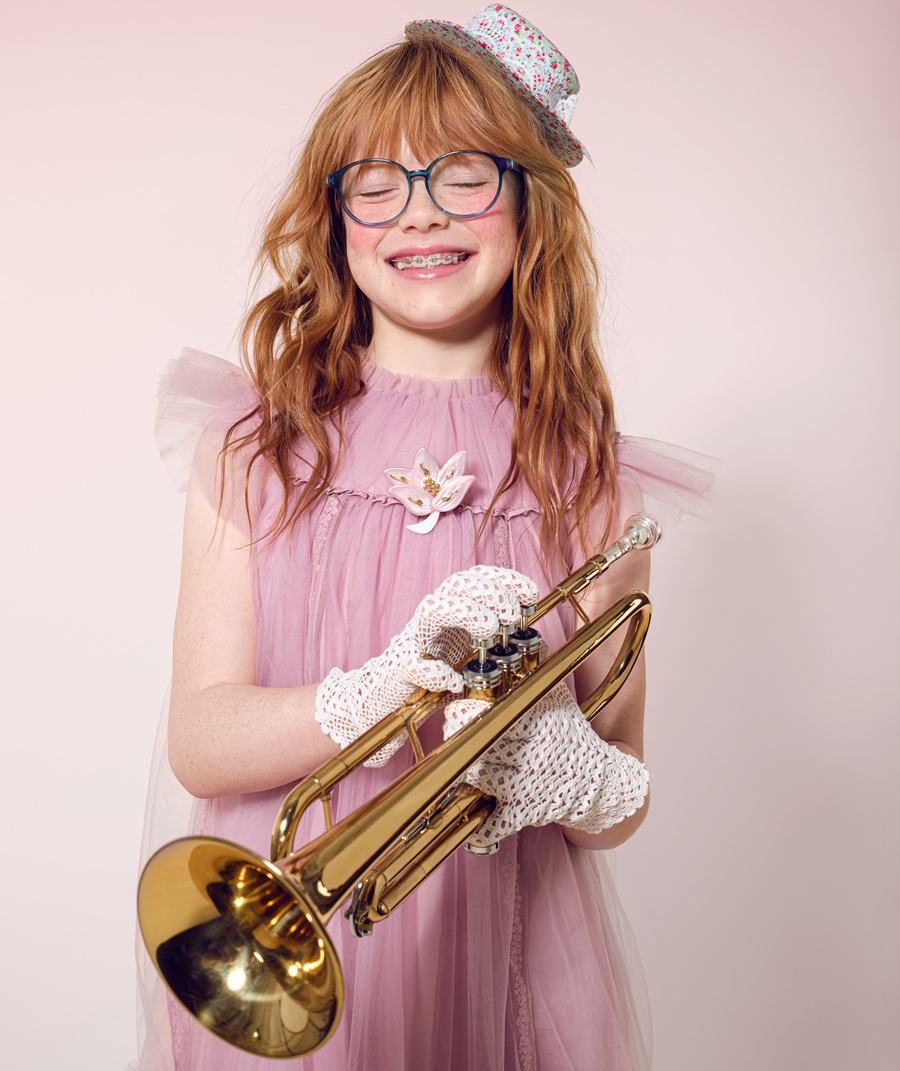 A red-haired girl in a pink tulle dress, lace gloves, a floral hat, and blue glasses beams with joy while holding a brass trumpet, embodying the spirit of a young musician lost in her own world.