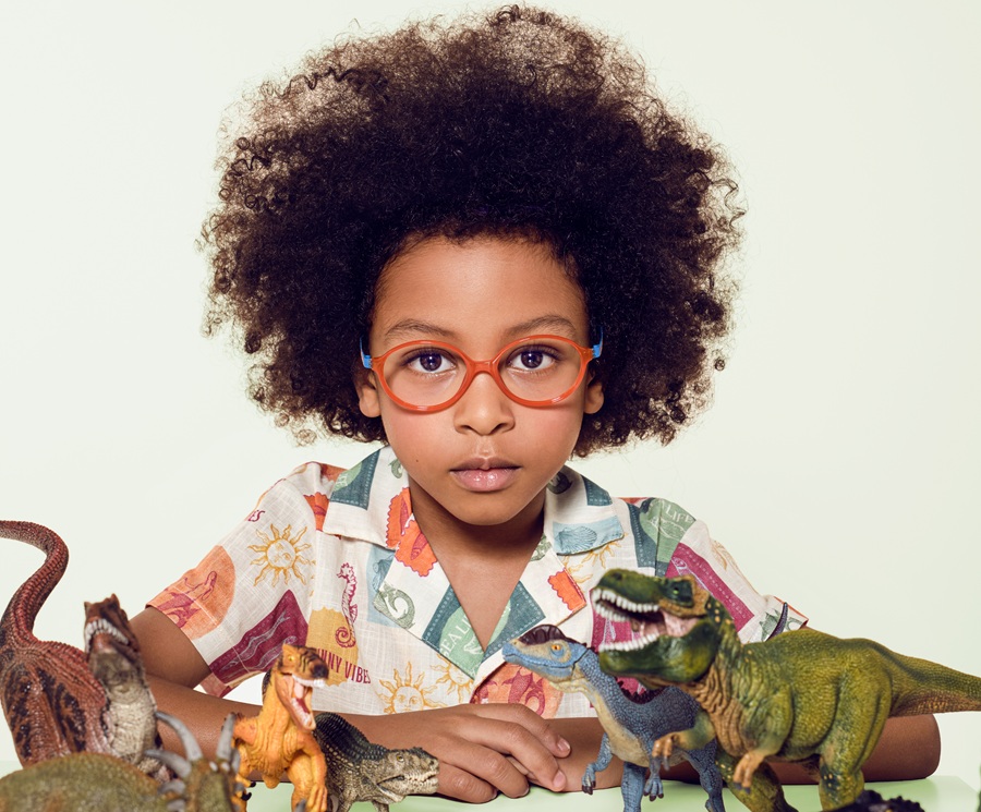 A young child with a large natural afro and orange-framed glasses rests their arms on a table surrounded by toy dinosaurs, gazing directly into the camera with quiet intensity. Part of the Lookkino 2026 campaign celebrating children's imagination.
