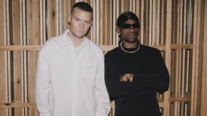 Matthew M. Williams et Travis Scott côte à côte devant un mur en lattes de bois. Williams porte une chemise blanche, Scott un pull noir avec des lunettes de soleil et un collier en chaîne.