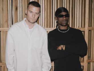 Matthew M. Williams et Travis Scott côte à côte devant un mur en lattes de bois. Williams porte une chemise blanche, Scott un pull noir avec des lunettes de soleil et un collier en chaîne.