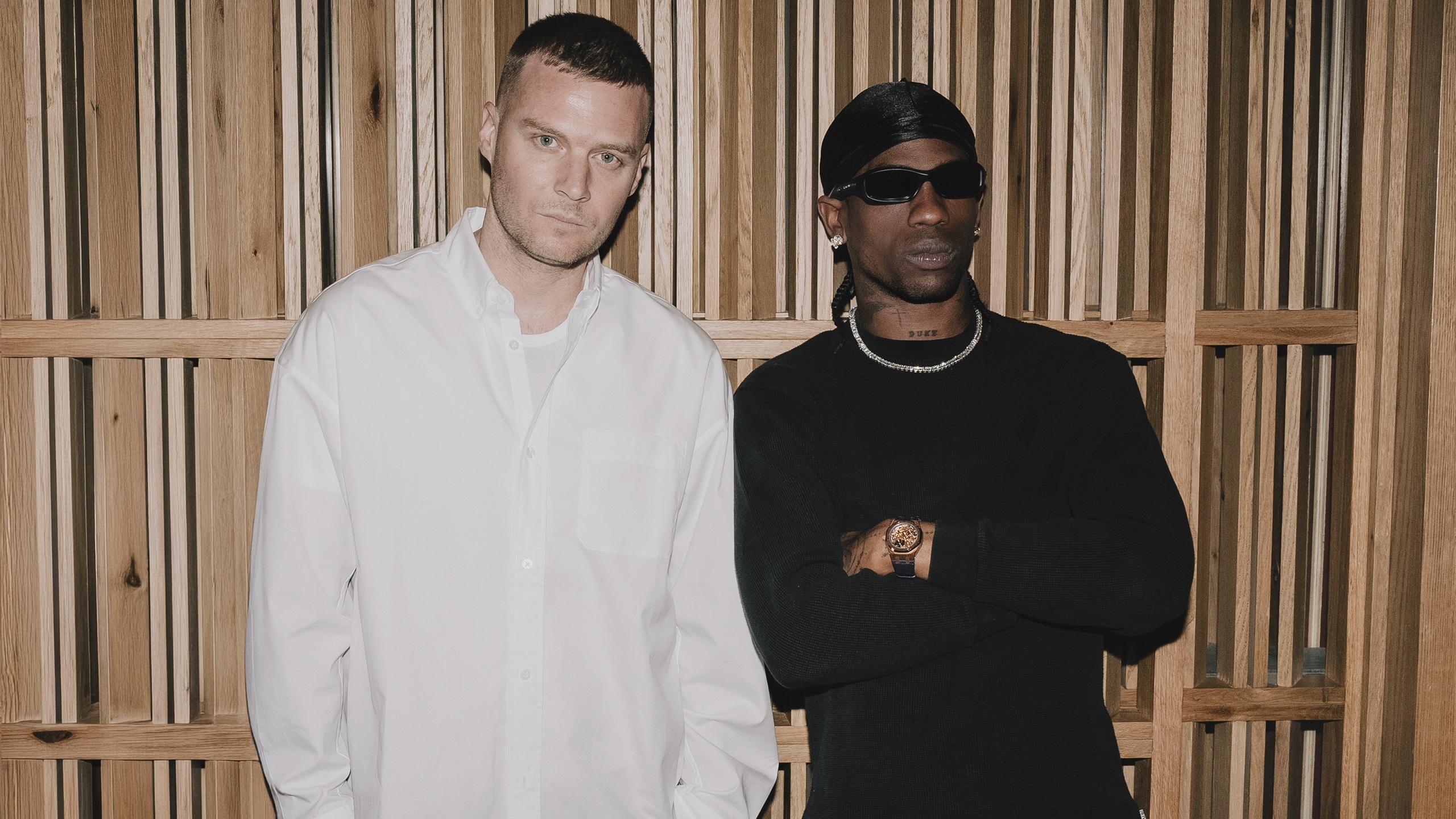Matthew M. Williams and Travis Scott standing side by side against a wooden slat wall. Williams wears a white shirt, Scott wears a black sweater with sunglasses and a chain necklace.