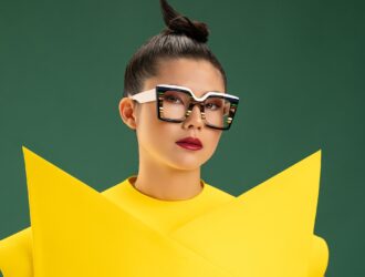 Model wearing Ultra Limited Acireale square optical frames in a layered tortoise and white acetate with multicolour stripe detailing, styled with a bold yellow structured top against a deep green background.