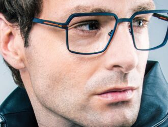 Un modèle masculin portant des lunettes J.F. Rey série SLEDGE en bleu marine mat avec des accents métalliques orange sur la charnière Flex. La grande monture géométrique carrée en métal est présentée dans un portrait en gros plan aux trois quarts, associée à une veste en cuir noir sur fond gris neutre. L'image dégage une esthétique architecturale et affirmée, fidèle à l'esprit de la collection SLEDGE alliant précision technique et innovation design.
