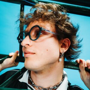 Young man wearing Spectaful FLARE optical frames in petrol green with grey acetate temples, photographed outdoors against a blue sky