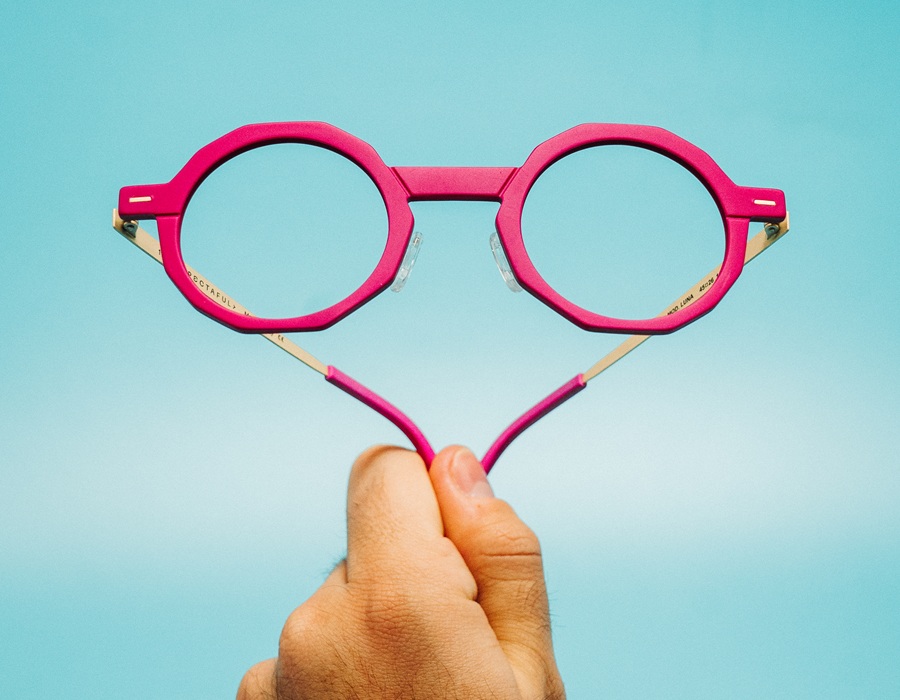 Monture optique Spectaful OBSIDIAN en façade polymère fuchsia tenue devant un fond bleu clair, branches ouvertes révélant un détail acétate doré