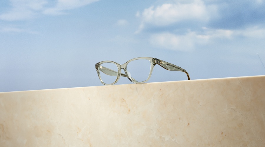 Montures optiques Tory Burch Kira œil-de-chat en gris translucide, posées sur un rebord en marbre sous un ciel bleu
