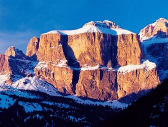 Snow-capped Dolomite rock formations glowing amber in the alpenglow light, with snow-dusted pine forests in the foreground and a deep blue winter sky above.