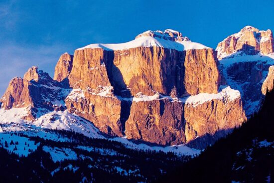 Snow-capped Dolomite rock formations glowing amber in the alpenglow light, with snow-dusted pine forests in the foreground and a deep blue winter sky above.