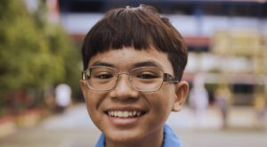 A young boy wearing glasses and a blue shirt smiles brightly at the camera, with a blurred school courtyard in the background.