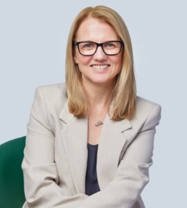 Jane Hoban, Managing Director of Specsavers Canada, seated and smiling, wearing black-rimmed glasses and a beige blazer.