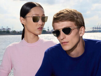 Two models wearing Oliver Peoples x Jil Sander titanium sunglasses from the debut collaboration. The woman wears a square flat-top metal frame; the man wears an angular half-rim style. Campaign photographed by Walter Pfeiffer in Hamburg.