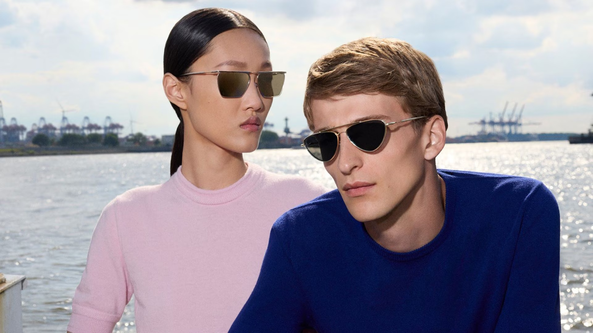 Two models wearing Oliver Peoples x Jil Sander titanium sunglasses from the debut collaboration. The woman wears a square flat-top metal frame; the man wears an angular half-rim style. Campaign photographed by Walter Pfeiffer in Hamburg.