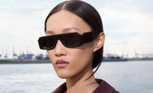A model wearing the Oliver Peoples x Jil Sander Edition 4 sunglasses — a bold acetate frame with an ultra-horizontal silhouette that creates a striking stripe across the face, shown in black. Shot by Walter Pfeiffer in Hamburg, with a waterfront and industrial cranes in the background.