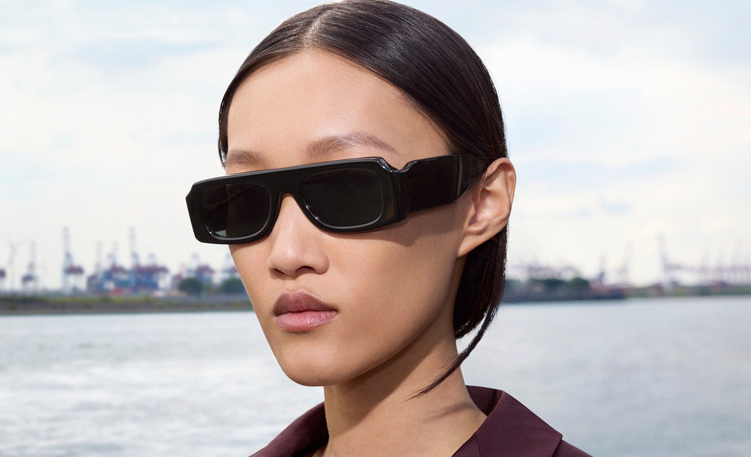 A model wearing the Oliver Peoples x Jil Sander Edition 4 sunglasses — a bold acetate frame with an ultra-horizontal silhouette that creates a striking stripe across the face, shown in black. Shot by Walter Pfeiffer in Hamburg, with a waterfront and industrial cranes in the background.