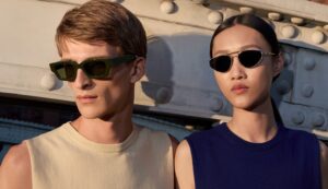 Deux mannequins portant des lunettes de soleil Oliver Peoples x Jil Sander dans un cadre industriel baigné d'une lumière dorée. L'homme porte une monture acétate rectangulaire Vert Foncé ; la femme porte une monture en titane à la géométrie épurée. Extrait de la campagne de la première collection, photographiée par Walter Pfeiffer.