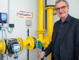 Thomas Windischbauer, COO of Silhouette Group, smiling next to industrial yellow gas pipes. He is holding a green lever on a gas valve, symbolically phasing out fossil fuel use at the Linz production site. A sign on the wall reads "Gashaupthahn" (Main Gas Tap).