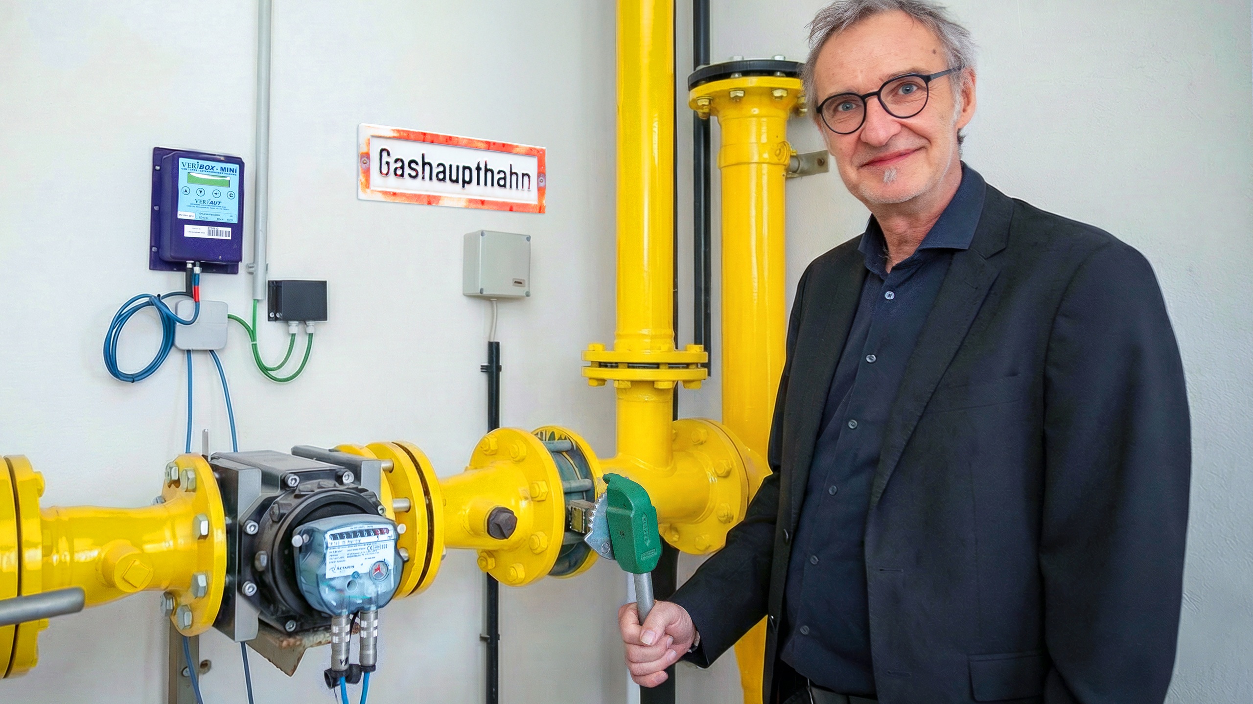 Thomas Windischbauer, COO of Silhouette Group, smiling next to industrial yellow gas pipes. He is holding a green lever on a gas valve, symbolically phasing out fossil fuel use at the Linz production site. A sign on the wall reads "Gashaupthahn" (Main Gas Tap).
