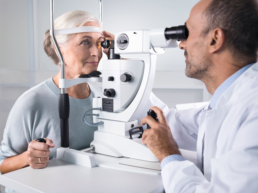An eye care professional using a ZEISS VISULAS green therapeutic laser to examine an elderly female patient at a slit lamp, in a bright clinical examination room.