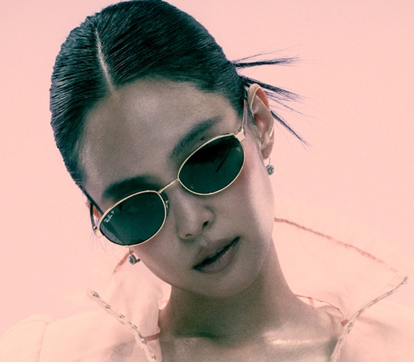 L'ambassadrice mondiale de Ray-Ban, Jennie, portant des lunettes de soleil Ray-Ban ovales à monture dorée et verres vert foncé, sur fond rose poudré dans la campagne de la marque