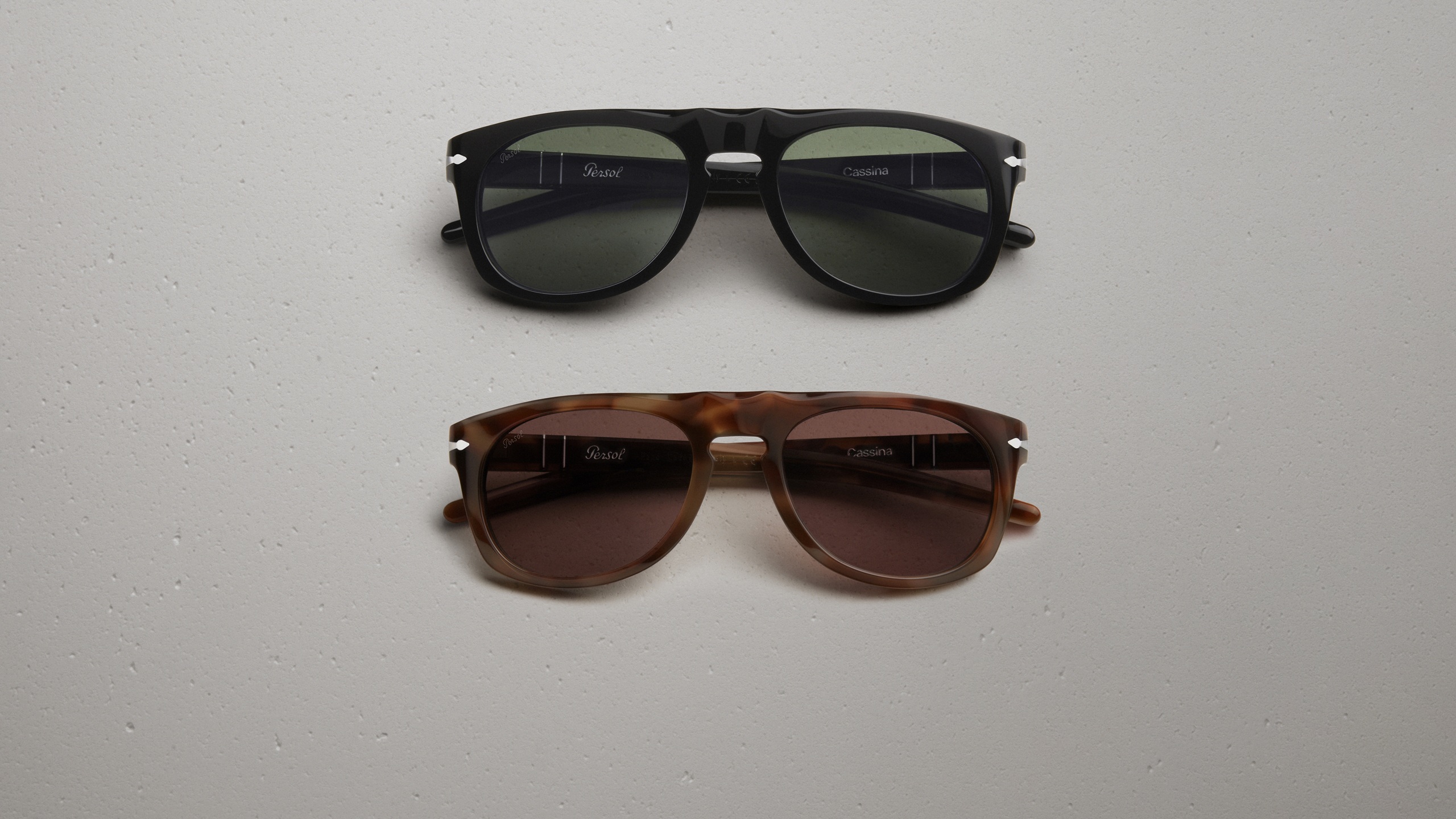 Lunettes de soleil Persol x Cassina en deux coloris : monture en acétate noir avec verres verts (en haut) et monture écaille havane avec verres marron (en bas), toutes deux arborant les logos Persol et Cassina sur la branche intérieure, posées à plat sur une surface texturée claire.