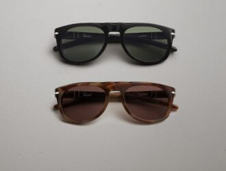 Persol x Cassina sunglasses in two colourways: a black acetate frame with green lenses (top) and a havana tortoiseshell frame with brown lenses (bottom), both featuring the Persol and Cassina logos on the inner temples, laid flat on a light textured surface.