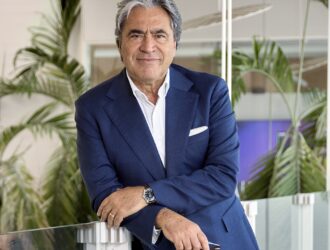 Angelo Trocchia, CEO of Safilo Group, wearing a navy blue suit and white dress shirt, leaning against a glass railing in a modern office setting with tropical plants in the background.