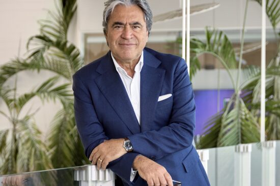Angelo Trocchia, CEO of Safilo Group, wearing a navy blue suit and white dress shirt, leaning against a glass railing in a modern office setting with tropical plants in the background.