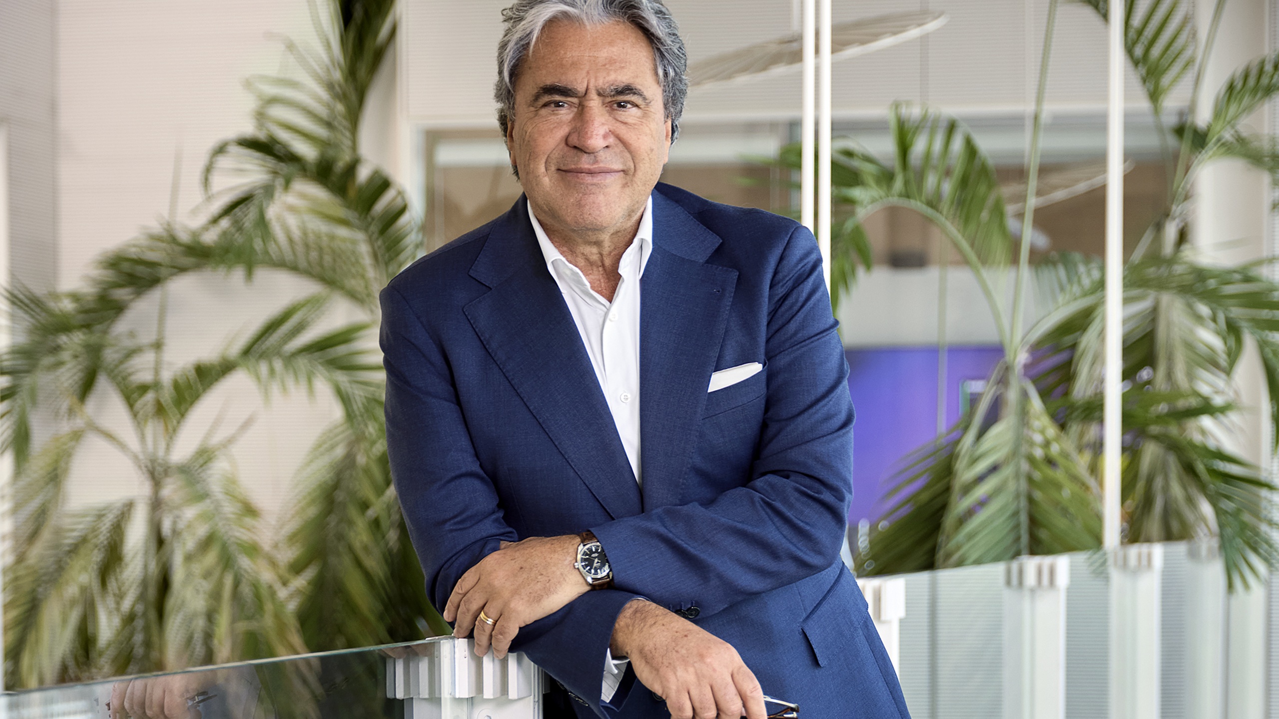 Angelo Trocchia, CEO of Safilo Group, wearing a navy blue suit and white dress shirt, leaning against a glass railing in a modern office setting with tropical plants in the background.