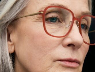 Close-up of a silver-haired woman wearing large square acetate eyeglass frames in translucent terra cotta red with slim gold metal temples, photographed against a dark background.