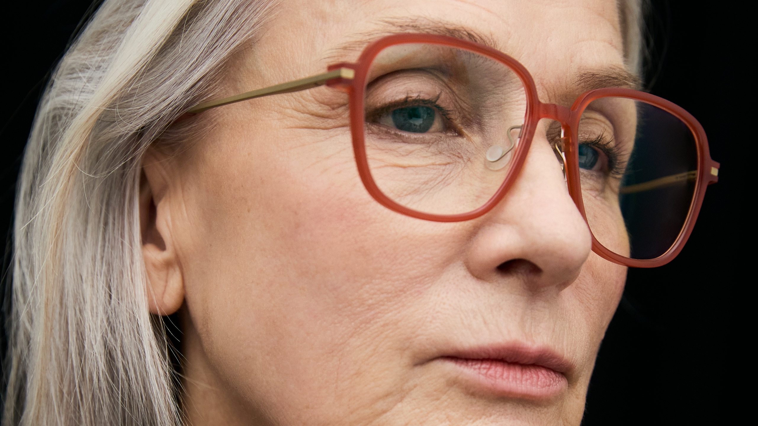 Close-up of a silver-haired woman wearing large square acetate eyeglass frames in translucent terra cotta red with slim gold metal temples, photographed against a dark background.